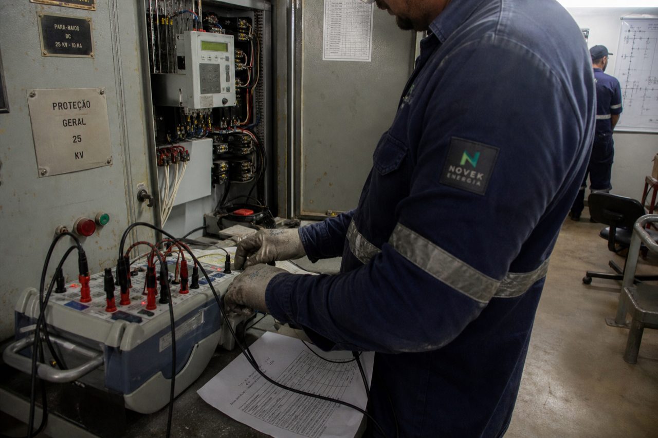 Técnico da Novek testando relé de proteção em cubículo de média tensão com equipamento de ensaio e cabos conectados